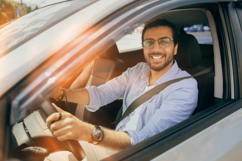 Handsome stylish man driving nice auto, looking through car window and smiling at camera